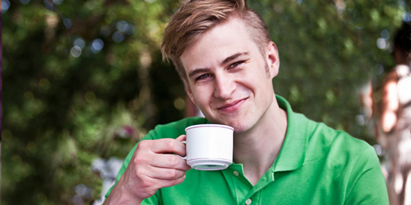 Frontalfoto: junger Mann in den 20ern, grünes Poloshirt, Kaffeetasse in der rechten Hand, freundlich lächelnd