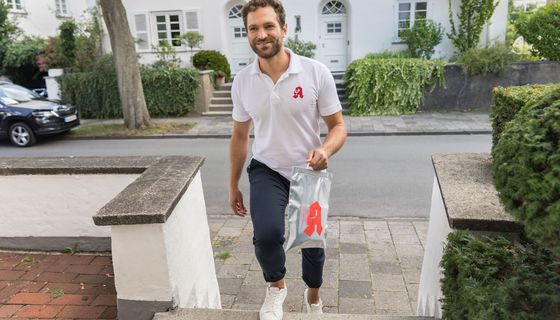 Mann mit Apothekenzeichen auf dem T-Shirt geht mit einer Arzneimitteltüte auf einen Hauseingang zu.