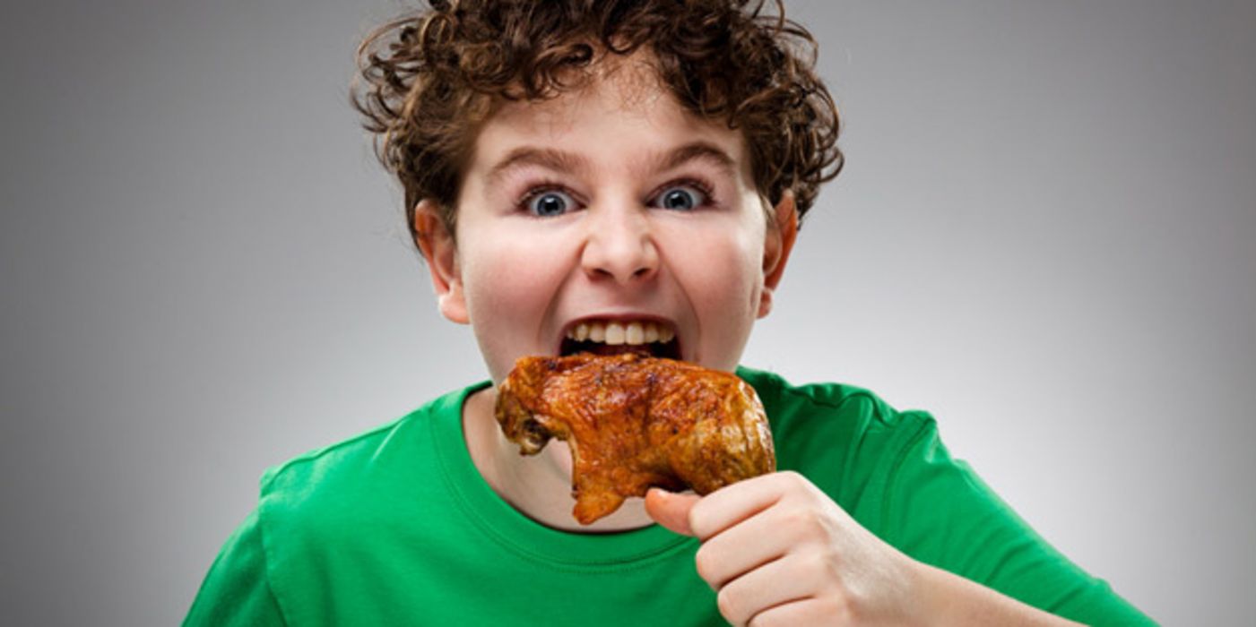 Frontalfoto eines Jungen mit dunklen Locken und grünem T-Shirt, der mit aufgerissenen Augen an einer Hühnerkeule nagt