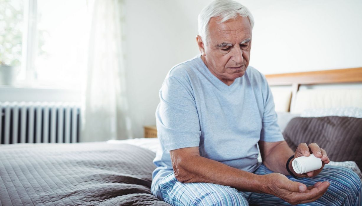 Mann, sitzt auf seinem Bett und schüttet eine Tablette aus einer Dose in seine Hand.