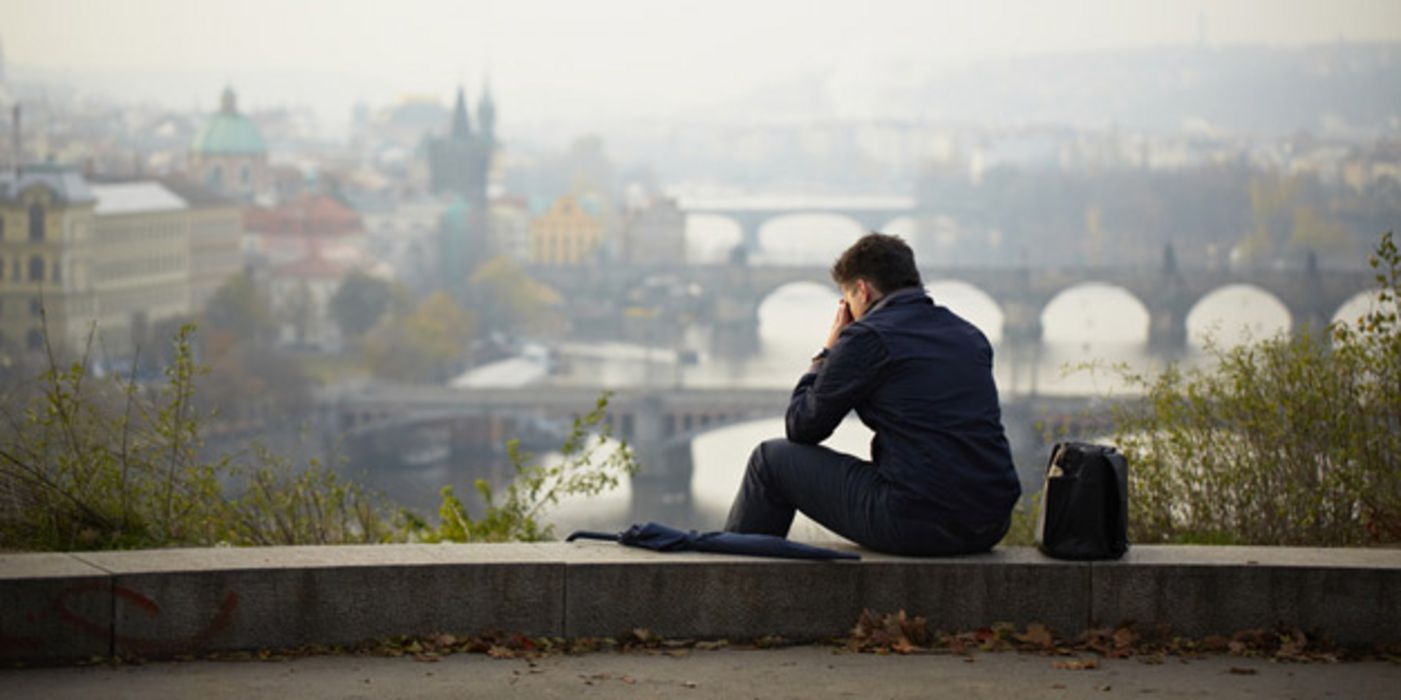 Rückansicht mit Halbprofil eines Mannes, auf einem Mäuerchen sitzend, das Gesicht in die Hände gestützt. Unter ihm die verhangene Stadtansicht von Prag und der Moldau