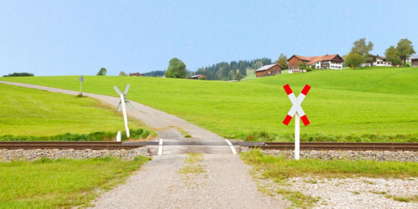 Bahnübergang auf dem Land, ein, zwei Häuser im Hintergrund, drumherum Wiesen