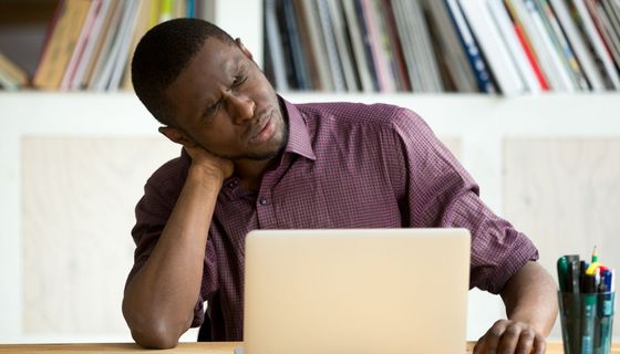 Mann am Laptopt mit sehr ordentlichem Schreibtisch greift sich in den Nacken und kneift die Augen zu.