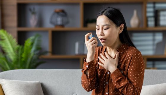 Frau sitzt auf dem Sofa und verwendet einen Inhalator. Eine Hand liegt an ihrem Schlüsselbein.