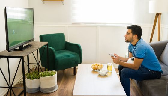 Junger Mann schaut ein Fußballspiel auf dem Fernseher. Auf dem Schoß hat er einen Fußball. Tageslicht fällt durch ein Fenster.