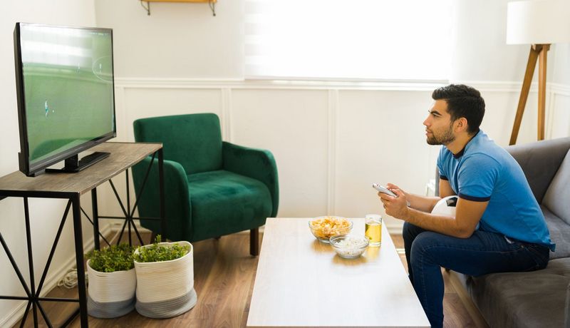 Junger Mann schaut ein Fußballspiel auf dem Fernseher. Auf dem Schoß hat er einen Fußball. Tageslicht fällt durch ein Fenster.