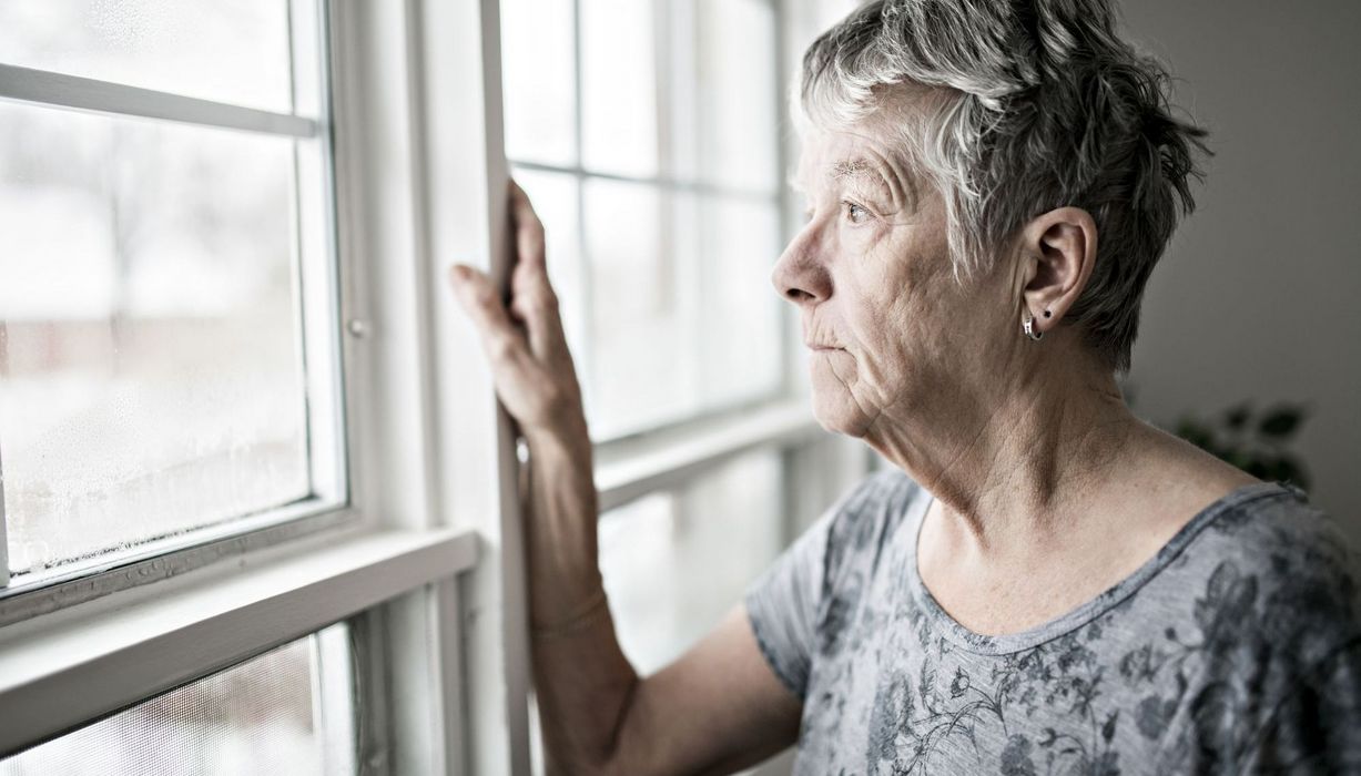 Ältere Dame, steht am Fenster und schaut traurig nach draußen.