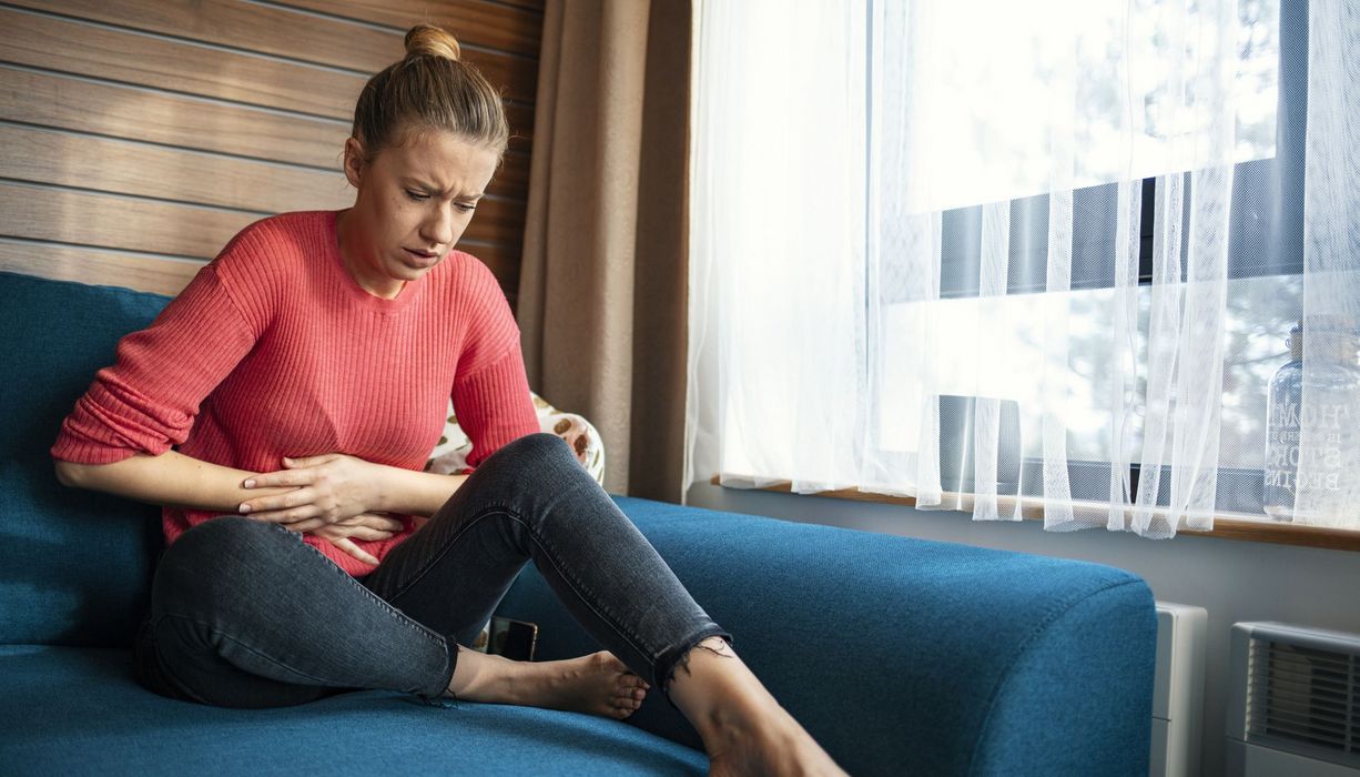 Junge Frau, sitzt auf der Couch und hält die Hände am Unterleib.