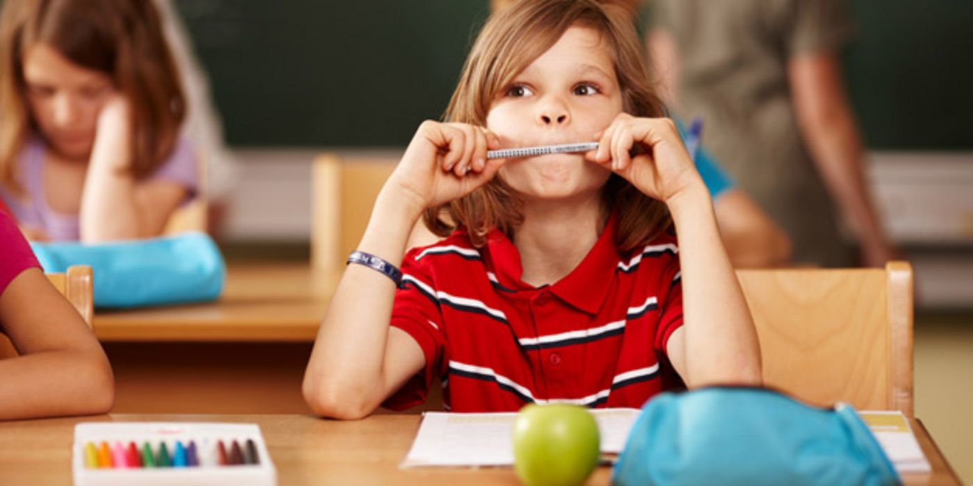 Grundschüler sitzt am Pult, hat einen Bleistift zwischen den Lippen und denkt nach