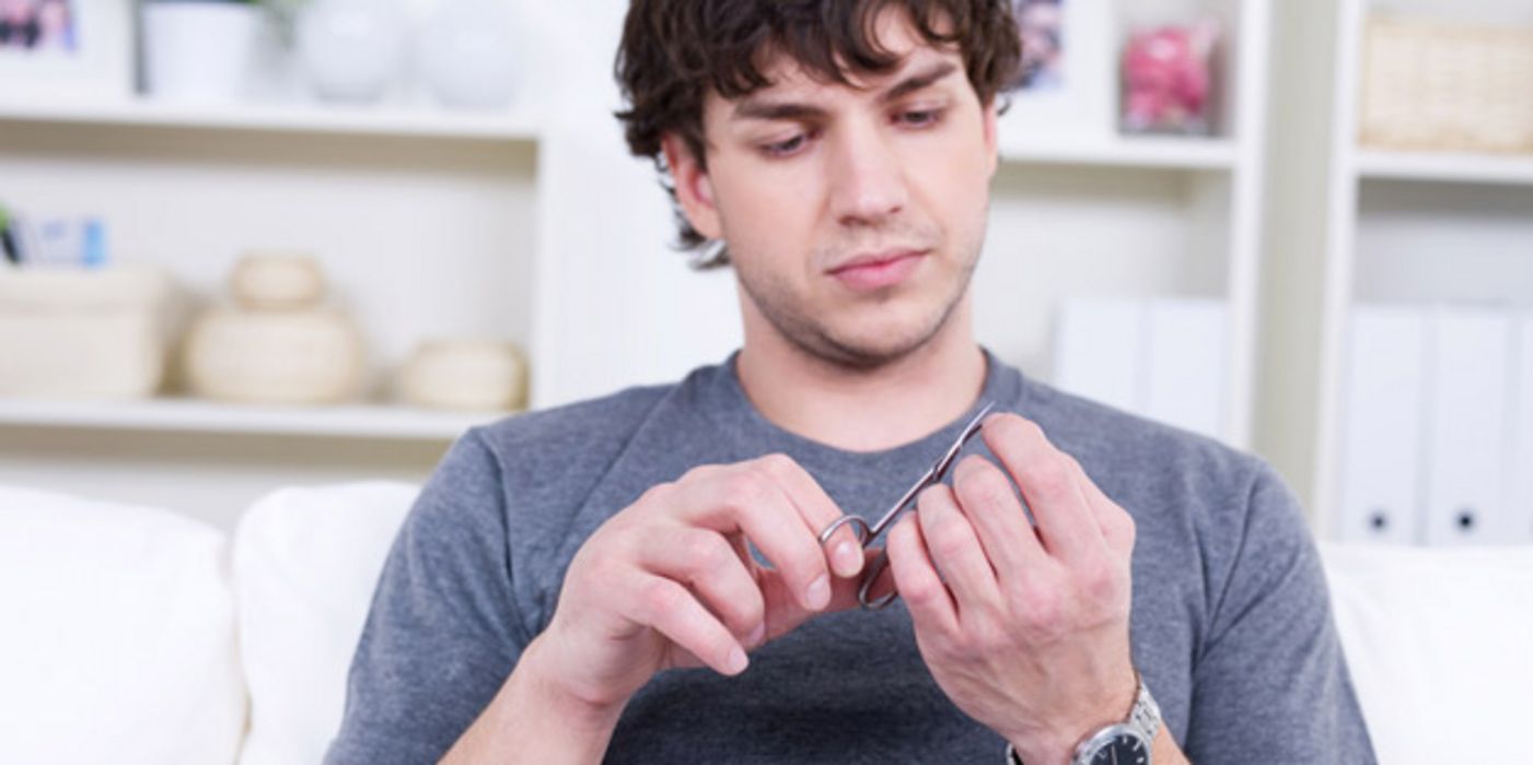 Brustbild: Mann in den 20ern, sportlich, graublaues T-Shirt, dunkles, lockiges Haar, schneidet sich konzentriert mit einer Nagelschere die Fingernägel