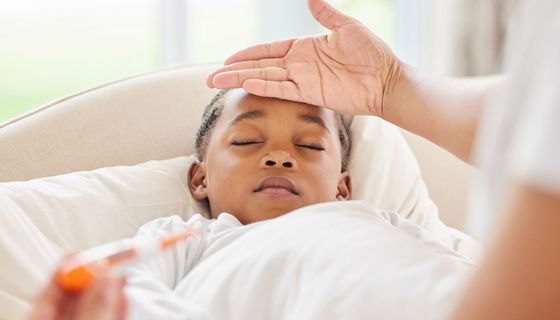 Krankes, schlafendes Kind im Bett, im Vordergrund (unscharf) ein Fieberthermometer, jemand hat dem Kind die Hand auf die Stirn gelegt.