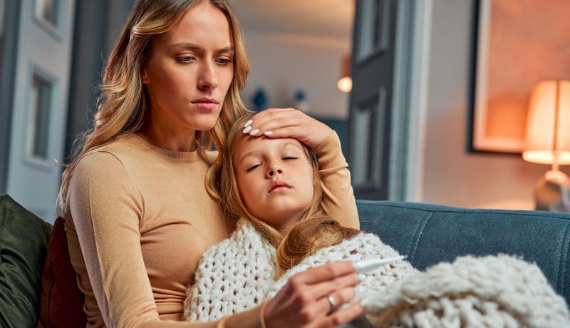 Krankes Kind liegt im Arm einer Frau, eingehüllt in eine Decke. Die Frau hat eine Hand an die Stirn des Kindes gelegt und liest ein Fieberthermometer ab.