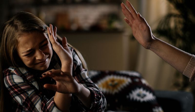 Mädchen duckt sich und hebt abwehrend die Hände – am rechten Bildrand ist eine drohend erhobene Hand zu sehen.