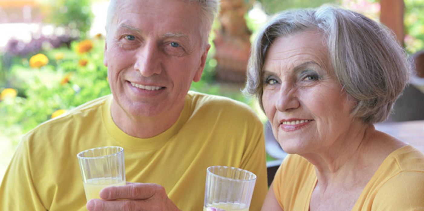 Sommerszene: Paar in den 60ern, Portrait, in die Kamera schauend, gelbe T-Shirts, jeweils ein Glas Limo in der Hand. Beide lächeln