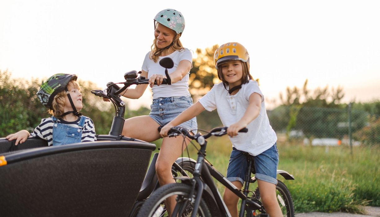 Junge Mutter mit Lastenrad und einem Kind darin, daneben ein weiteres Kind auf dem Fahrrad.