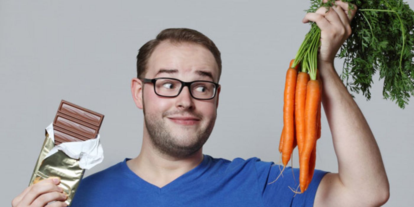 Junger Mann mit blauem T-Shirt hat in der einen Hand ein Bund Möhren, in der anderen eine Tafel Schokolade