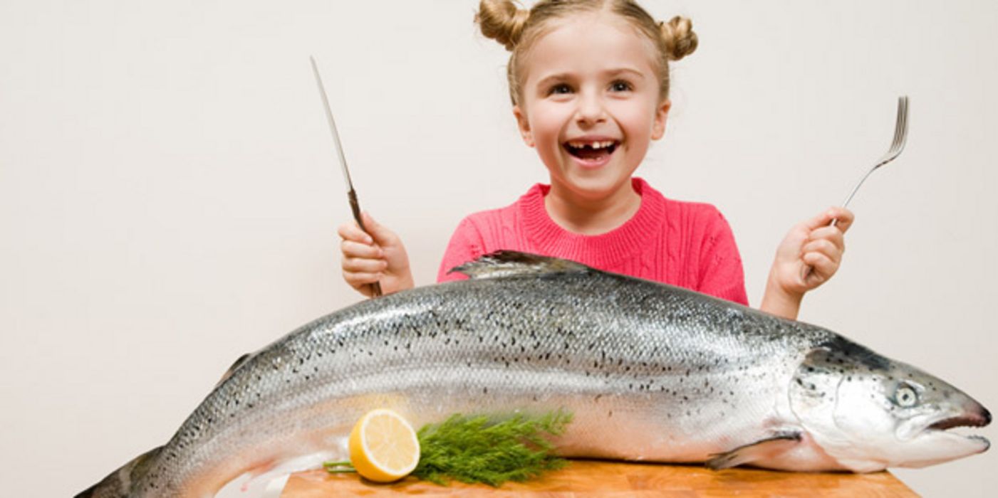 Blondes Mädchen lachend mit Messer und Gabel, auf dem Tisch vor sich einen großen Lachs