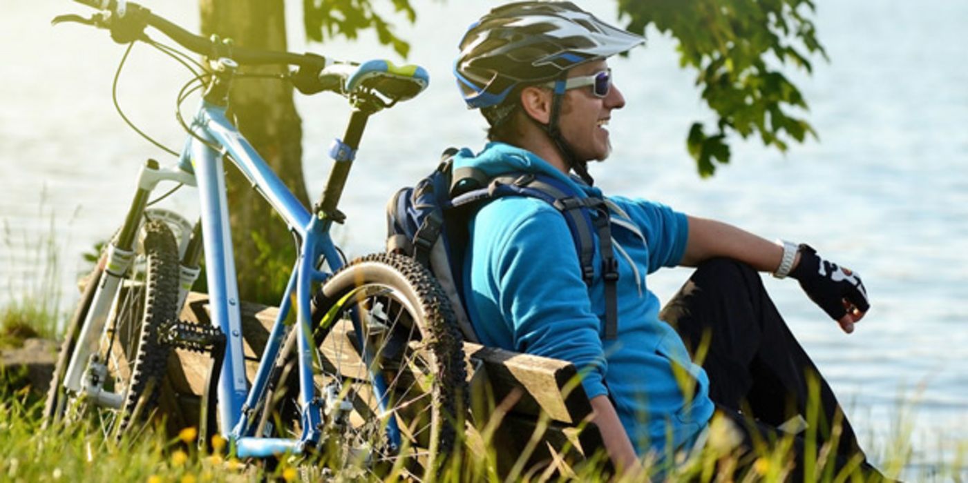 Mann um die 50 in Fahrradkleidung mit Helm lehnt an einem See am Baum, Mountainbike an die andere Seite des Baumes gelehnt. Abendstimmung