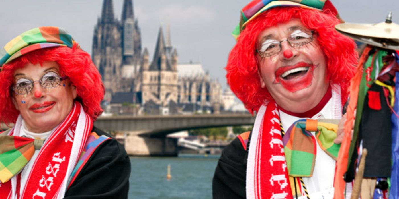 Karnevalistisches Paar in Clowsnsschminke und Hüten mit roten Perücken lackend vor Rhein und Kölner Dom