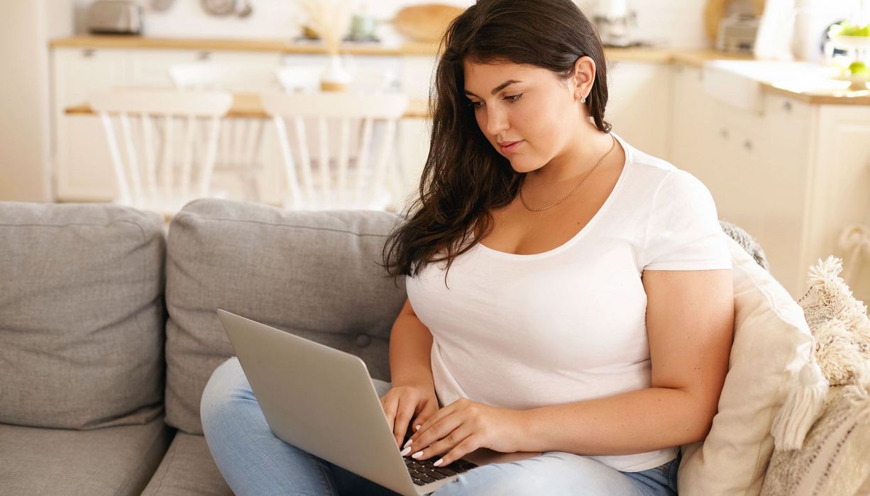 Übergewichtige Junge Frau, sitzt auf dem Sofa, Laptop auf dem Schoß.