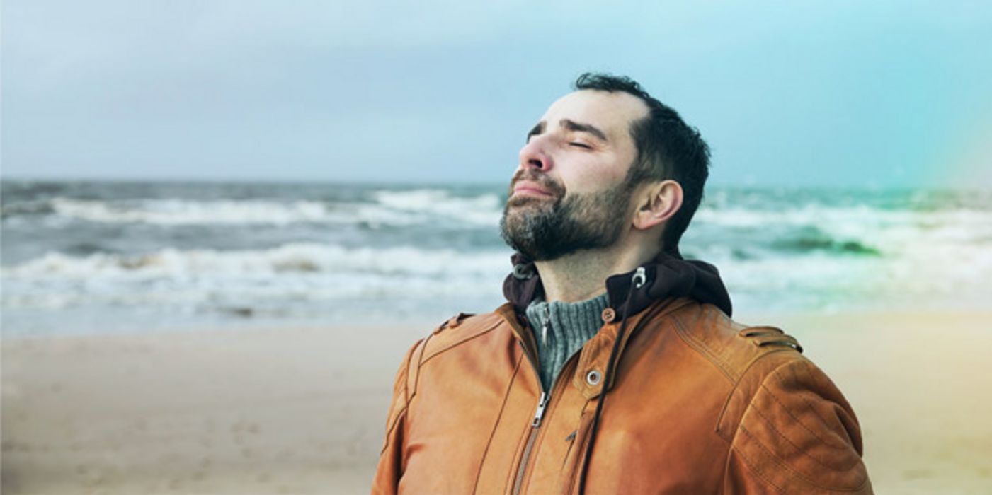 Mann am Meer (Oberkörper), rehbraune Lederjacke, schwarze Haare, Vollbart, ca. Ende 30, atmet mit geschlossenen Augen tief ein