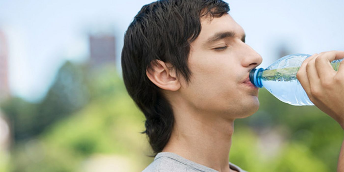 Junger Mann mit dunklen Haaren (Profilfoto vor unscharfem Naturhintergrund) trinkt aus einer Wasserflasche