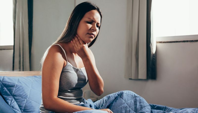 Junge Frau, sitzt im Bett und hat Halsschmerzen.