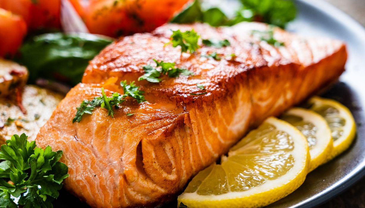 Auf einem Teller liegt Fisch, daneben Zitronenscheiben, Tomaten und grünes Gemüse. 