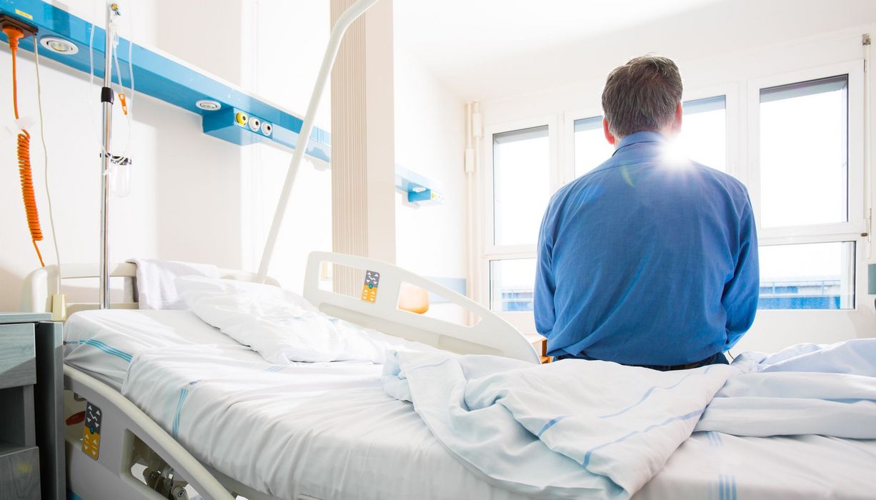 Ein Mann sitzt mit dem Rücken zum Betrachter auf einem Krankenhausbett.