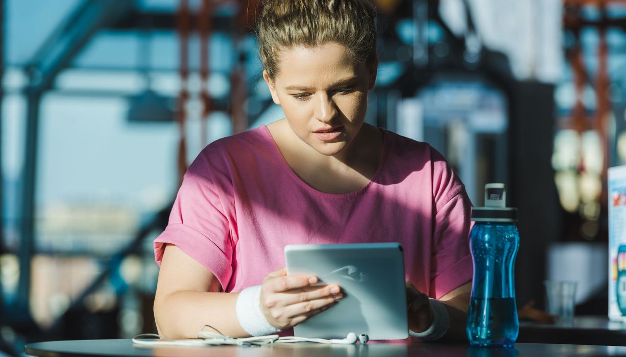 Frau mit Sportkleidung, sitzt vor ihrem Tablet mit einer Wasserflasche.