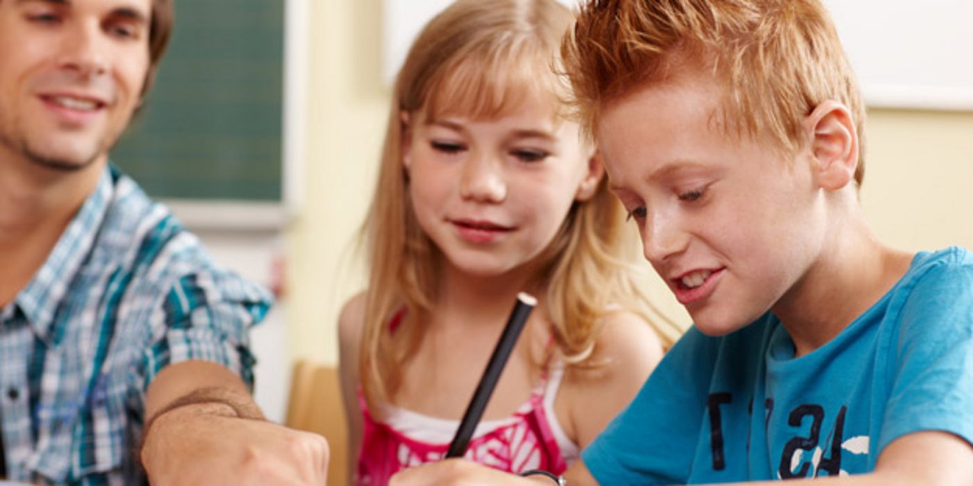 Ein Junge und ein Mädchen sitzen im Klassenraum und bekommen von einem jungen Lehrer etwas erklärt