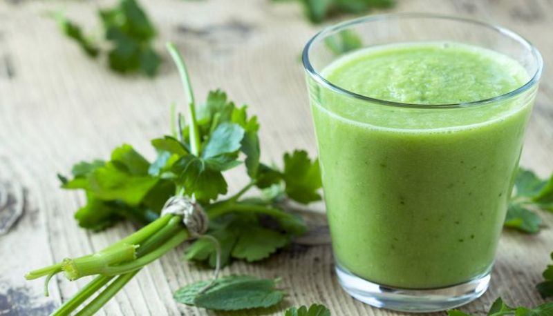 Glas mit grünem Smoothie auf Holztisch, drumherum grüne Kräuter