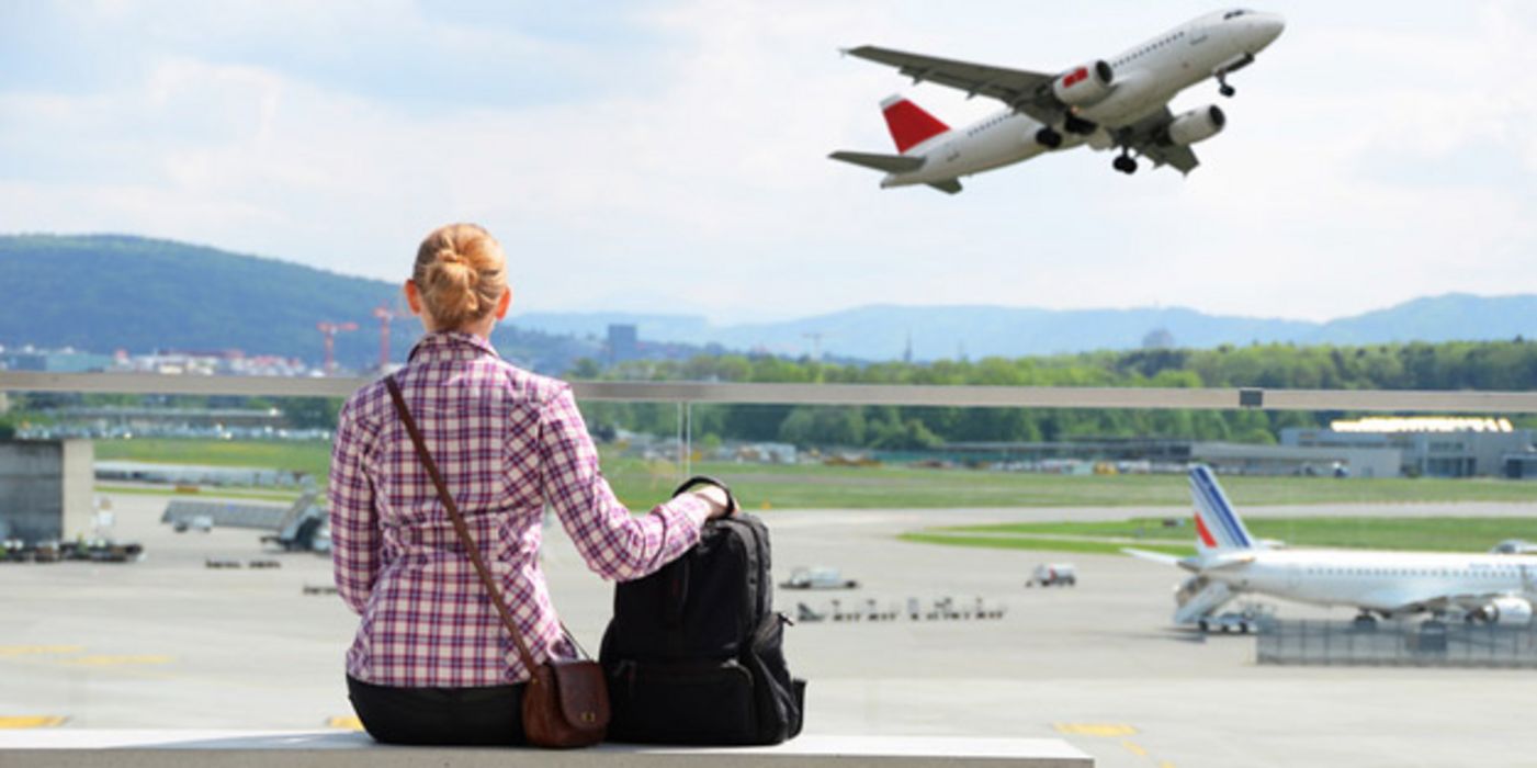 Rückansicht einer blonden Frau mit Fluggepäck in rotkarierter Bluse, die auf einer Bank sitzend den Abflug eines Flugzeuges beobachtet