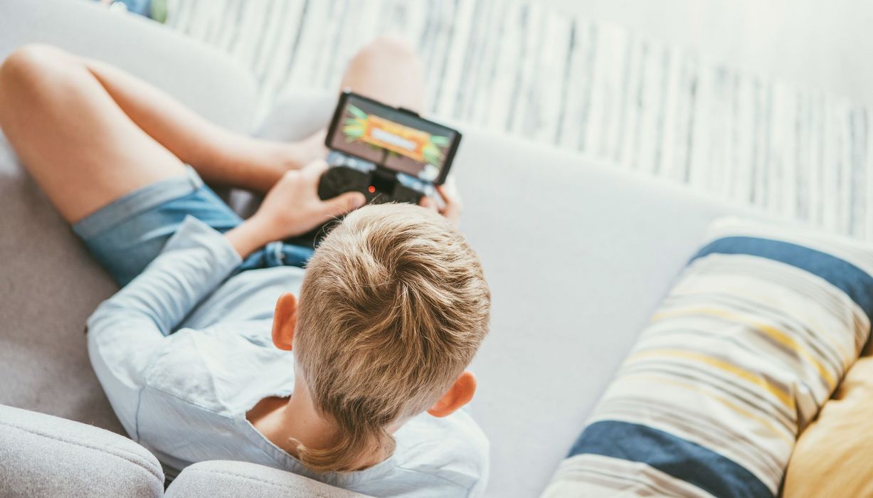 Blonder Junge, sitzt auf einer Couch und spielt mit einer Spielekonsole. 