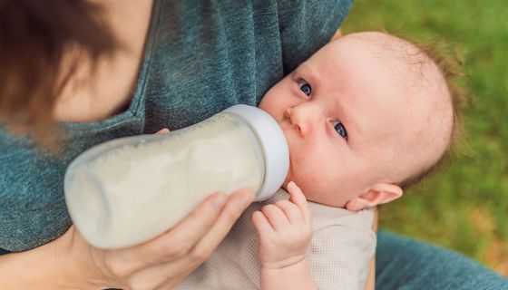 Baby trinkt auf dem Schoß einer Frau aus einer Flasche. Es blickt zweifelnd in die Kamera.