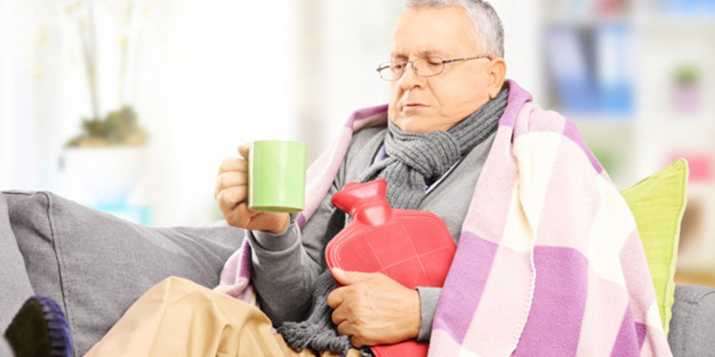 Grippekranker Mann in den 60ern auf der Couch mit bunter Decke, Schal, Wärmeflasche, Trinkbecher