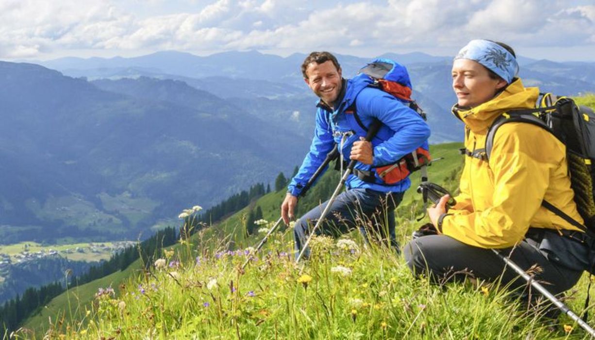 Mann und Frau auf einer Bergblumenwiese, Wanderanoraks, Rucksäcke, Blick in ein Tal, Sonnenschein, Mann lächelt Frau an