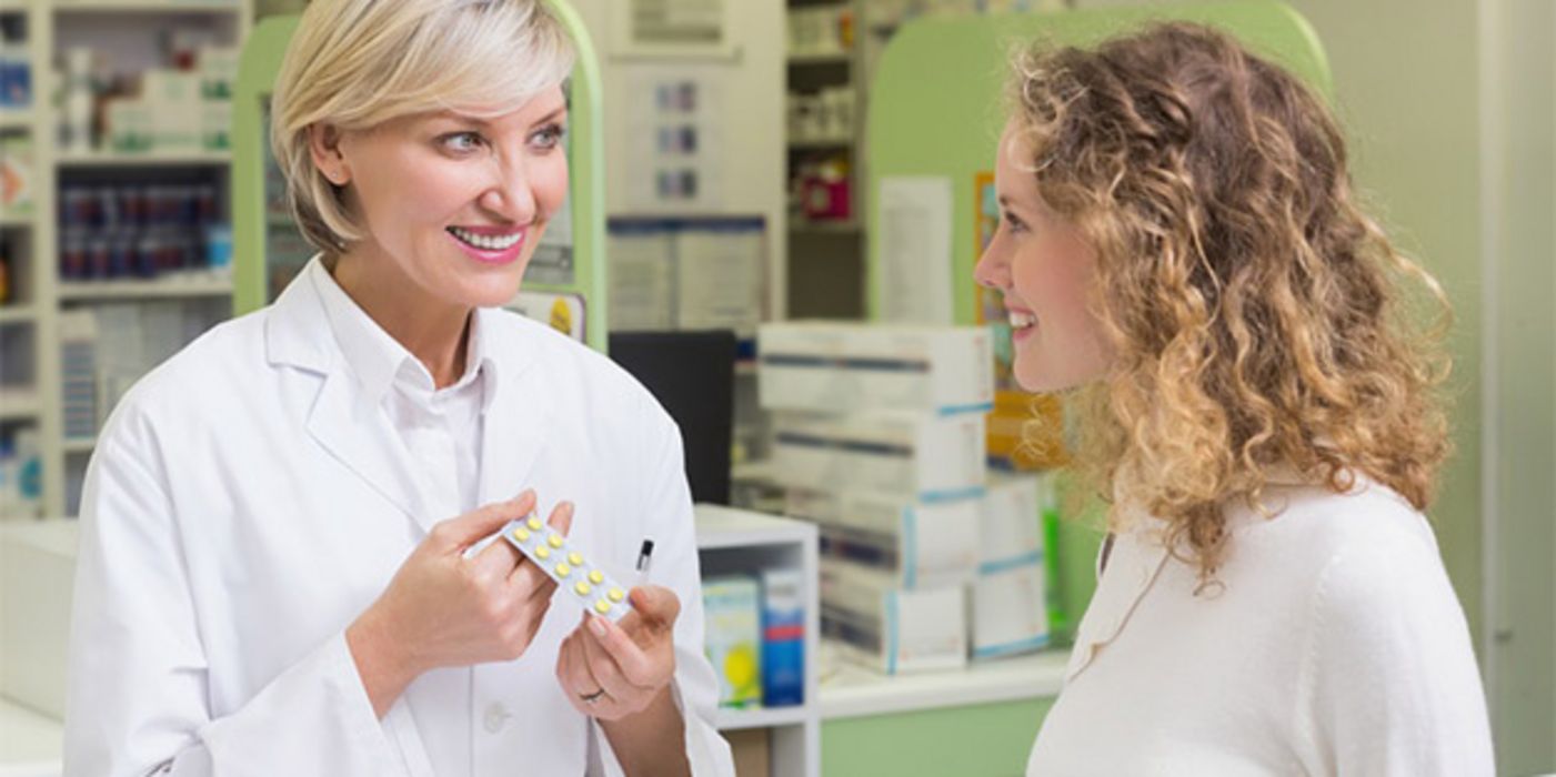 Beratungsgespräch: Apothekerin (in den 30ern, blond) mit Blister in der Hand berät Kundin in den 20ern (schulterlange, leicht gelockte, mittelblonde Haare). Beide lächeln