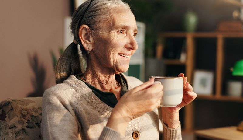Eine Frau über 65 hält lächelnd eine Tasse in beiden Händen.