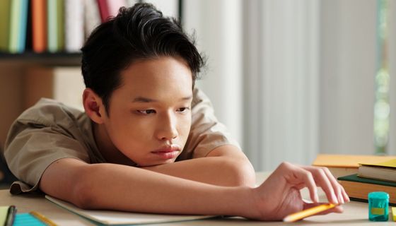 Junge am Schreibtisch liegt mit den Armen auf einem Heft. Er spielt mit einem Bleistift und einem Spitzer.