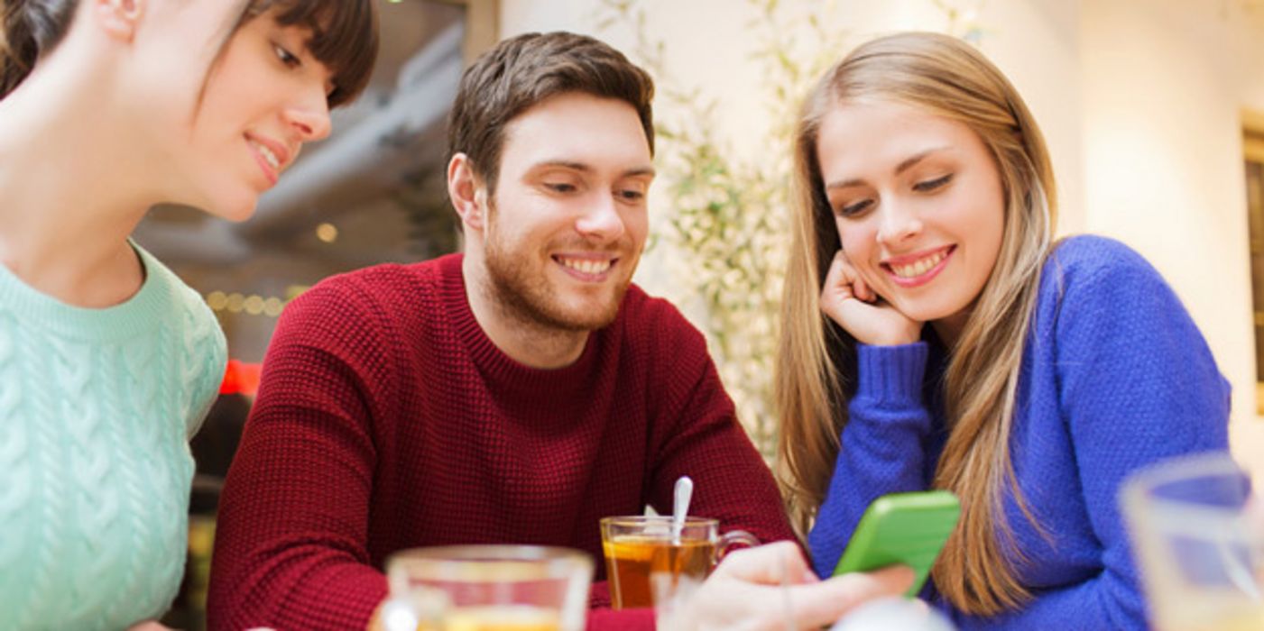 Junge Leute um die 20 (2 Frauen, 1 Mann), am Tisch sitzend, schauen lachend in grünes Smartphone, das der Mann in der Hand hält