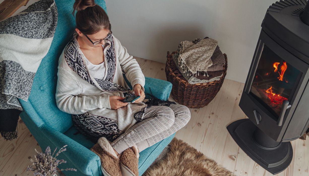 Junge Frau, sitzt im Winter auf einem Sessel vor dem Kamin, mit Handy in der Hand.