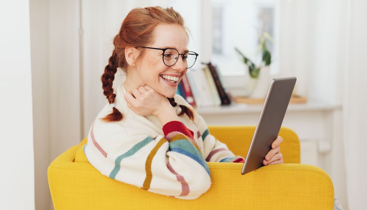 Junge Frau, sitzt auf der Couch und schaut lächelnd auf ihr Tablet.