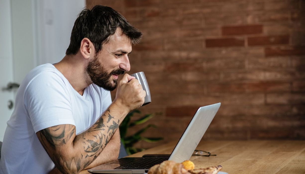 Mann, ca. 40 Jahre alt, sitzt am Schreibtisch vor seinem Laptop und trinkt Kaffee.