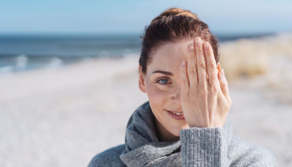 Frau am Strand, hält ein Auge mit einer Hand zu.