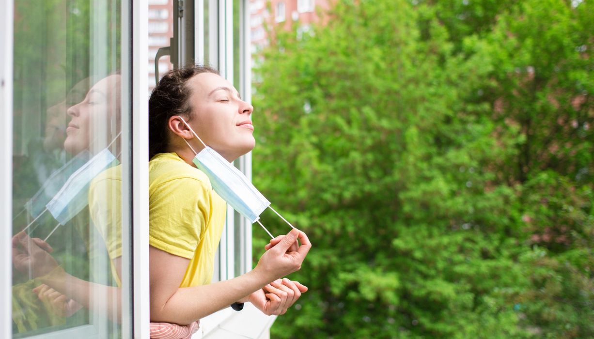 Junge Frau, lehnt sich aus dem Fenster und zieht Mund-Nasen-Schutz ab.