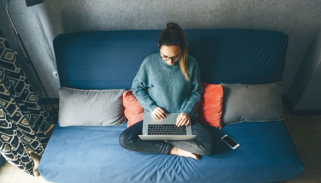 Junge Frau, sitzt im Schneidersitz auf der Couch, Laptop auf dem Schoß.