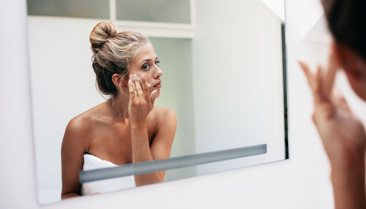 Junge blonde Frau, steht vor dem Spiegel und cremt sich das Gesicht ein.