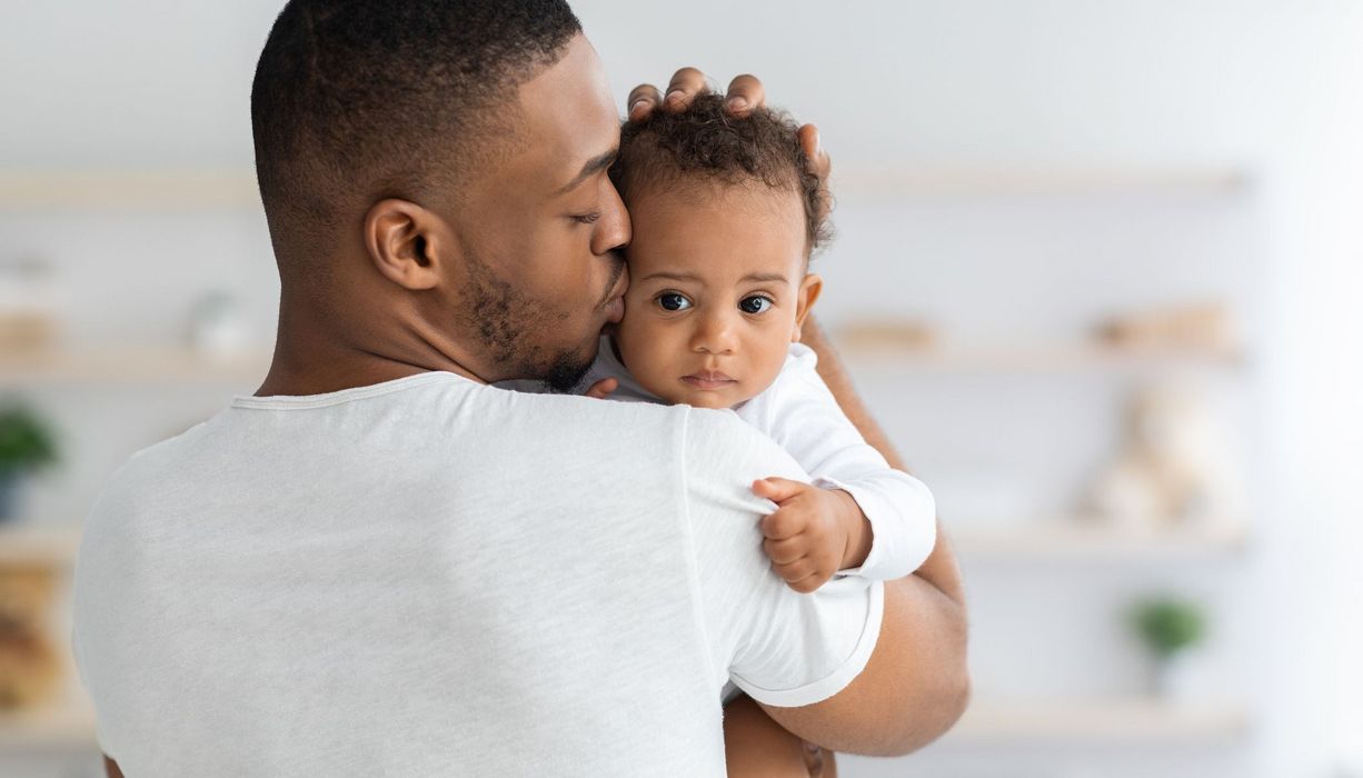 Vater, hält sein Baby auf dem Arm und küsst es auf die Stirn.