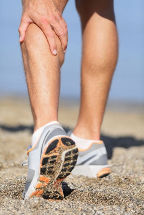 Mensch in Turnschuhen am Strand greift sich an die linke Wade.